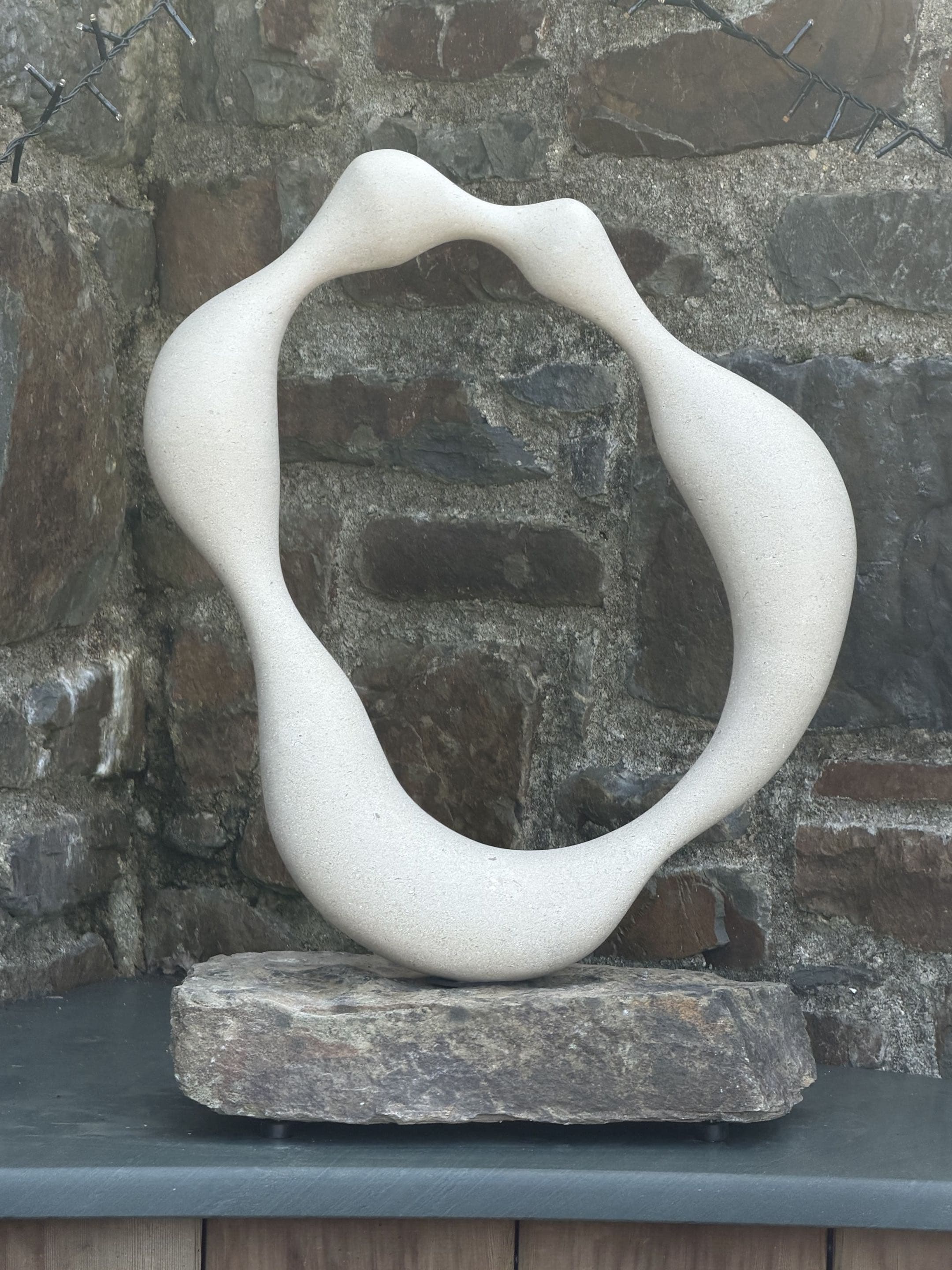 Pete Carling's stone sculpture 'The Kiss 4', a smooth carved loop form set on a stone plinth against a Devon stone wall